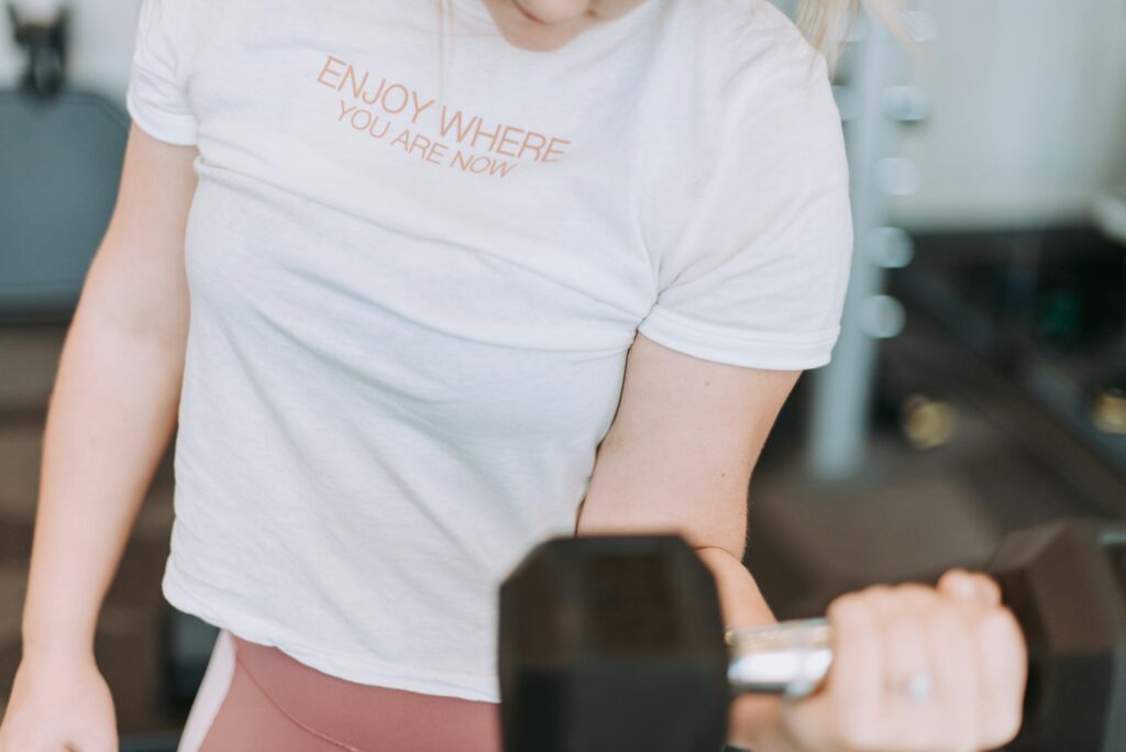 Torso of person working out, raising a dumb bell in a bicep curl. T-shirt reads, "Enjoy where you are now."